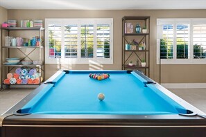 A pool table with a blue felt surface, surrounded by shelves with various items and a window with blinds. - Home Theater | Private Pool | Hot Tub | Mini Golf (Scottsdale)