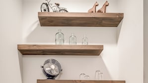 Una kitchenette compacta con microondas, un refrigerador pequeño y una cafetera sobre la encimera. Hay repisas de madera arriba que contienen varios objetos, incluyendo una bicicleta y adornos.