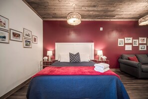 Une chambre à coucher avec un lit, une table de chevet, une lampe, un mur rouge et un canapé.
