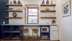 Une cuisine avec des étagères en bois, une fen être, deux tabourets, un four à micro-ondes et un réfrigérateur.