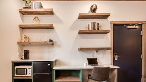 Una cocina compacta con microondas, refrigerador y un portátil sobre una encimera blanca. Hay estantes de madera arriba con libros y objetos decorativos. Se ve una puerta a la derecha.