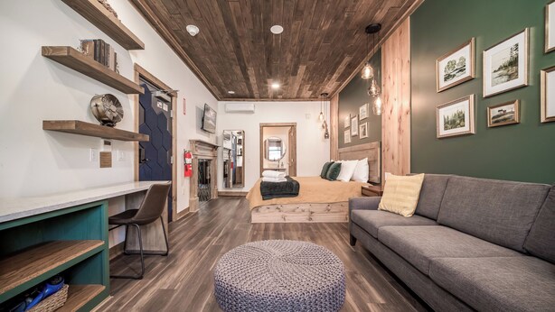 Una habitación de hotel moderna con techo de madera, un sofá gris, un otoman redondo y una cama con manta color beige.