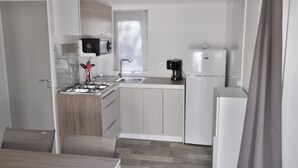 Een kleine keuken met een kookplaat, wastafel en koelkast, houten keukenkastjes en een raam met zonwering.