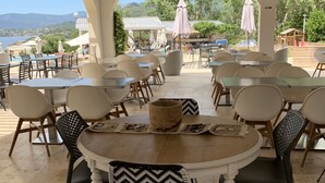 Een terras met ronde tafels en stoelen, een houten plafond en uitzicht op zee en heuvels.