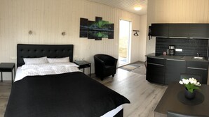 A modern hotel room with a large bed, a small desk, a chair, a kitchenette, and a view of the outdoors.