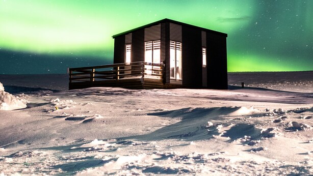 A small, dark wooden building with a wooden deck in front, situated on a snowy landscape under a night sky with visible stars and the aurora borealis.
