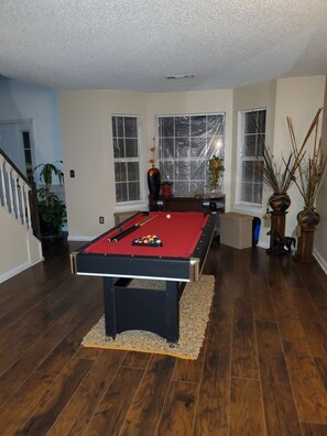 A pool table in a room with wooden flooring, large windows with blinds, and a staircase leading to another level.