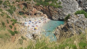 Ein felsiger Strand mit klarem, blauem Wasser, umgeben von spärlicher Vegetation und Klippen.