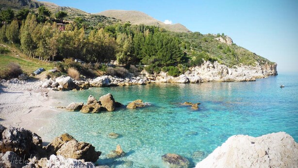 Eine Küstenlandschaft mit klarem türkisblauem Wasser, felsigen Ufern und einer Hügelslope mit dichtem Grün.