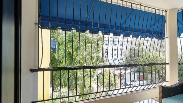 Un balcón con mobiliario de mimbre, una mesa de vidrio y toldos azules.