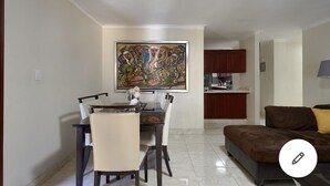 Un comedor con una mesa y sillas, un cuadro sobre la mesa, una cocina al fondo y un sofá al frente.
