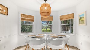 A dining room with a round glass table, white chairs, a woven pendant light, and windows with blinds. - Magnolia House- Walkable Uptown, Kids Playhouse, Peaceful Peacocks, Near Beach! (St. Augustine)