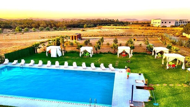 Una grande piscina circondata da comode sedie a sdraio bianche e tende, con vista su dolci colline e un edificio sullo sfondo.
