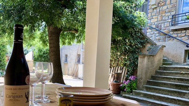 A bottle of Carignan wine, two wine glasses, and a can of beer on a wooden table with a view of a stone building and a staircase. - Gites Corbieres- rental with private pool, near Narbonne (Montséret)