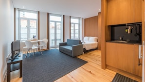 Habitación de hotel moderna con un ventanal grande, un sofá gris, una cama, un pequeño espacio de cocina y un escritorio con una silla.