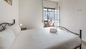 Un letto rifatto con un copriletto bianco e cuscini, una lampada da comodino e una finestra con vista sugli edifici.