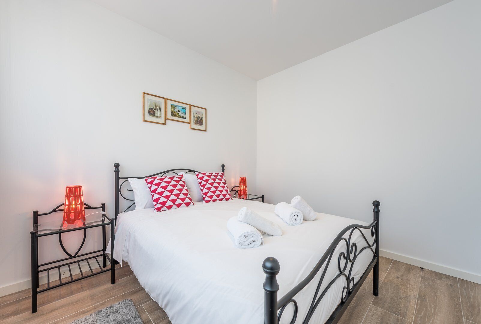 A neatly made bed with white bedding and red and white patterned pillows, a metal-framed bed frame, and two red lamps on either side.