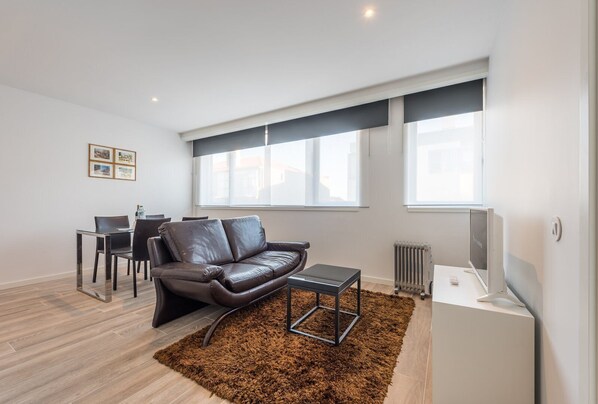 A modern living room with a leather sofa, a glass coffee table, and a small rug. There is a dining area with a table and chairs, and large windows providing natural light. - GuestReady - Retreat near Matosinhos beach (Matosinhos)