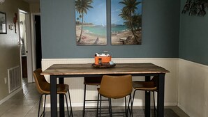 Ein Essbereich mit einem Holztisch, vier Stühlen, einer Deckenleuchte und zwei an der Wand befestigten Bildern einer Strandansicht.