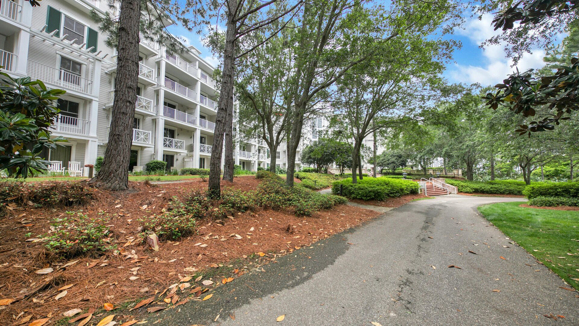 A pathway leading to a multi-story building with balconies, surrounded by trees and greenery.