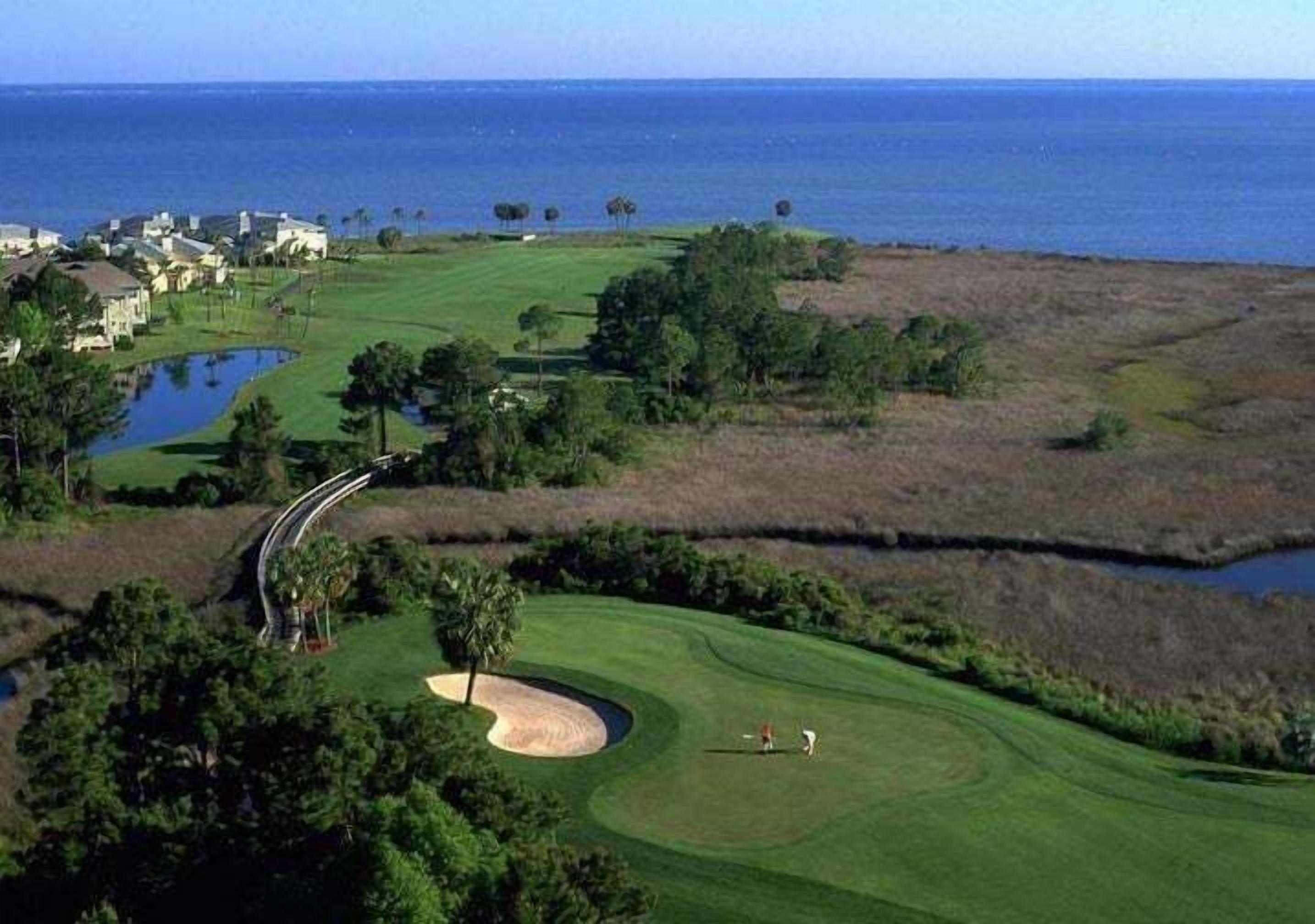 An aerial view of a golf course with a water body, trees, and a distant view of the ocean.