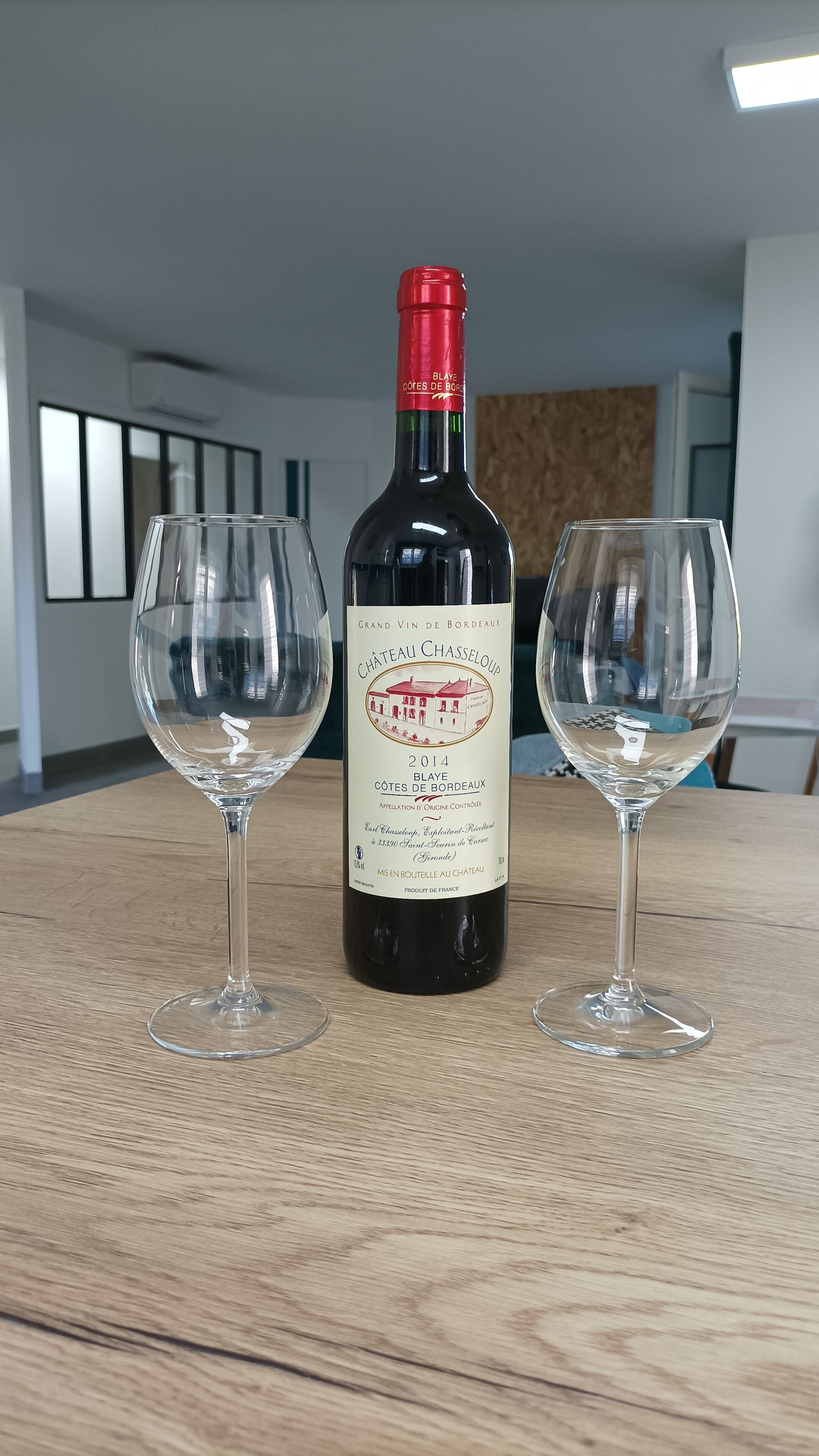 A bottle of Chateau Chasseloup wine with two empty wine glasses on a wooden table.
