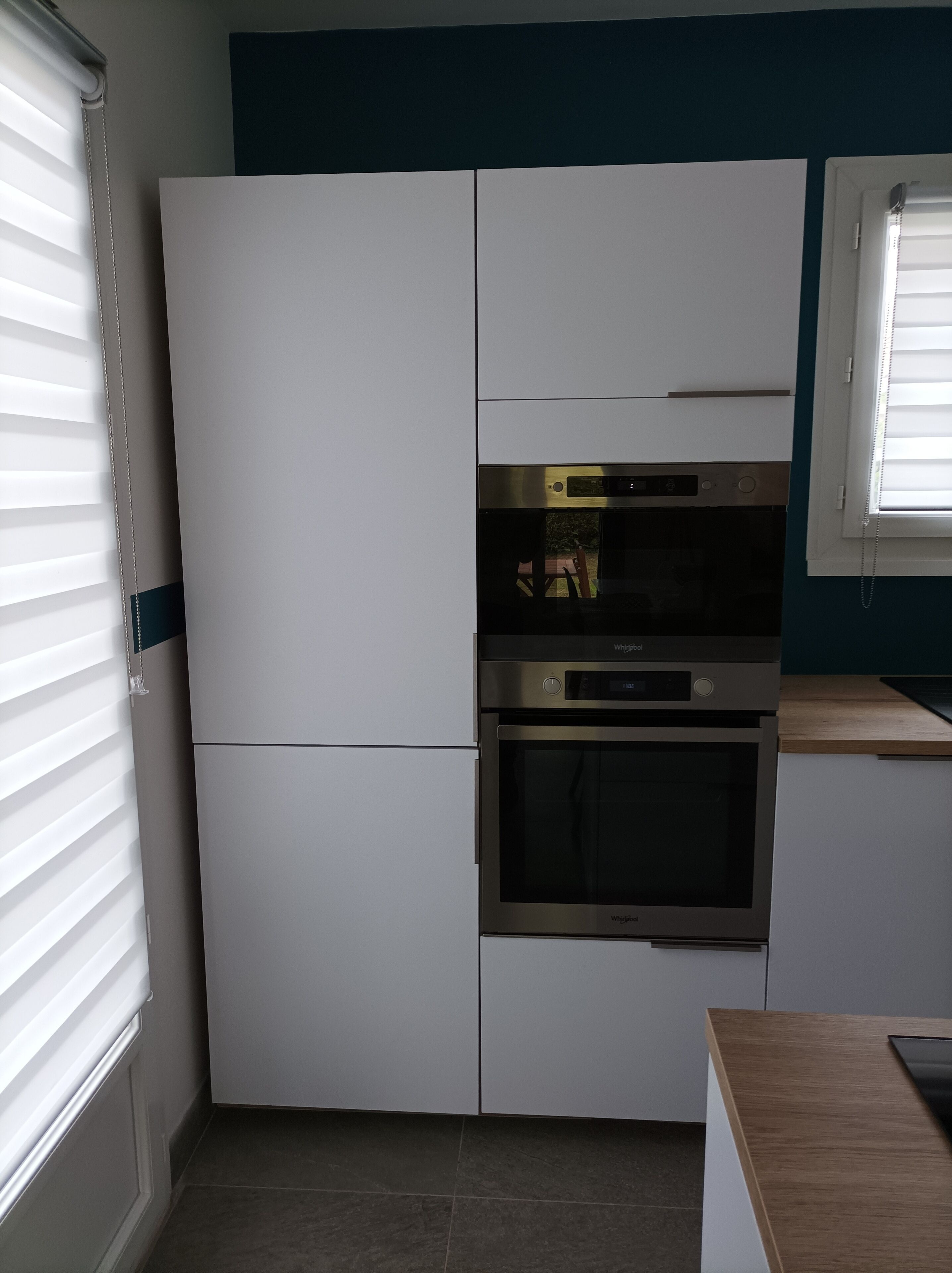 Una cucina compatta con forno integrato e microonde, mobili bianchi e una finestra con tende.