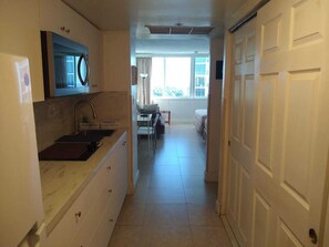 A narrow hallway leading to a bedroom with a window, a kitchen area with a microwave, and a door on the right.