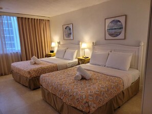 Quarto de hotel com duas camas, cada uma com colcha estampada e almofadas, uma mesa de cabeceira com abajur e uma janela com cortinas.