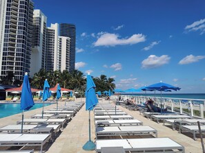 Área de piscina com espreguiçadeiras e guarda-sóis, céu claro e um edifício alto ao fundo.