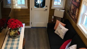 A cozy cabin interior with a wooden ceiling, a dining area with a checkered tablecloth, a black sofa with decorative pillows, and a small window with a pink umbrella. - ALL INCLUSIVE AMENITIES:Kayak,SUP,Pedal Boat,PingPong,Pickleball,Movie Nt &MORE! (Nice)