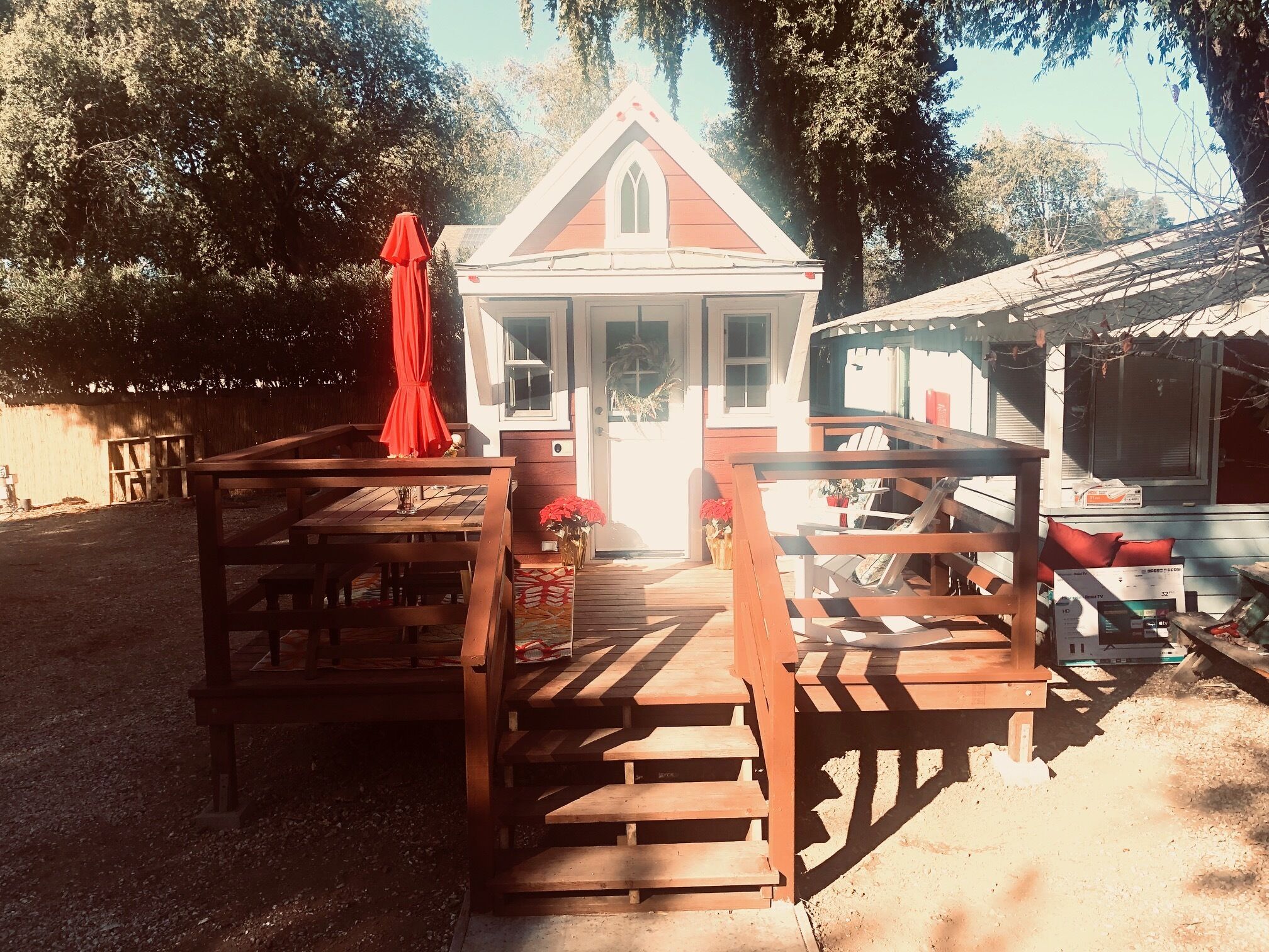 Una terraza de madera con una mesa, sillas, un paraguas rojo y un pequeño edificio con porche.
