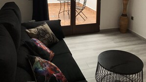 Een woonkamer met een zwarte bank, kleurrijke kussens, een kleine ronde tafel en een schuifdeur naar een terras buiten met een tafel en stoelen.