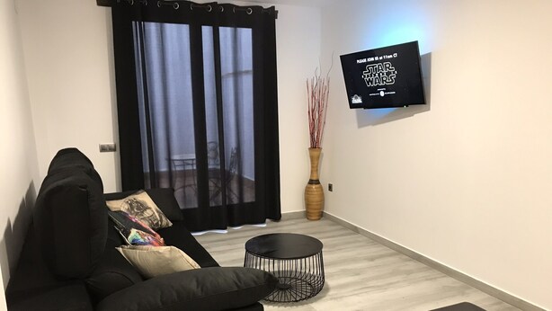 Een moderne woonkamer met een zwarte bank, een ronde zwarte salontafel en een flatscreen-televisie aan de muur met een Star Wars poster.