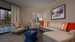 Una habitación de hotel con un ventanal grande, un sofá, dos sillones, un otoman azul y un cuadro en la pared.