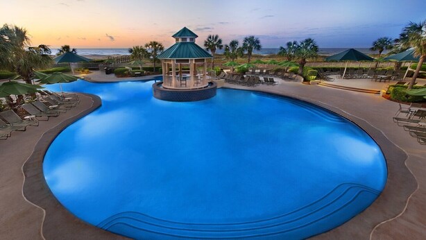 Una piscina grande y circular con un quiosco en el centro, rodeada de sillas de descanso y sombrillas, y vista al océano al atardecer.