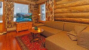 Interior de una cabaña de troncos con techo de madera, un televisor sobre un soporte de madera, un sofá, una mesita redonda pequeña y cortinas estampadas.