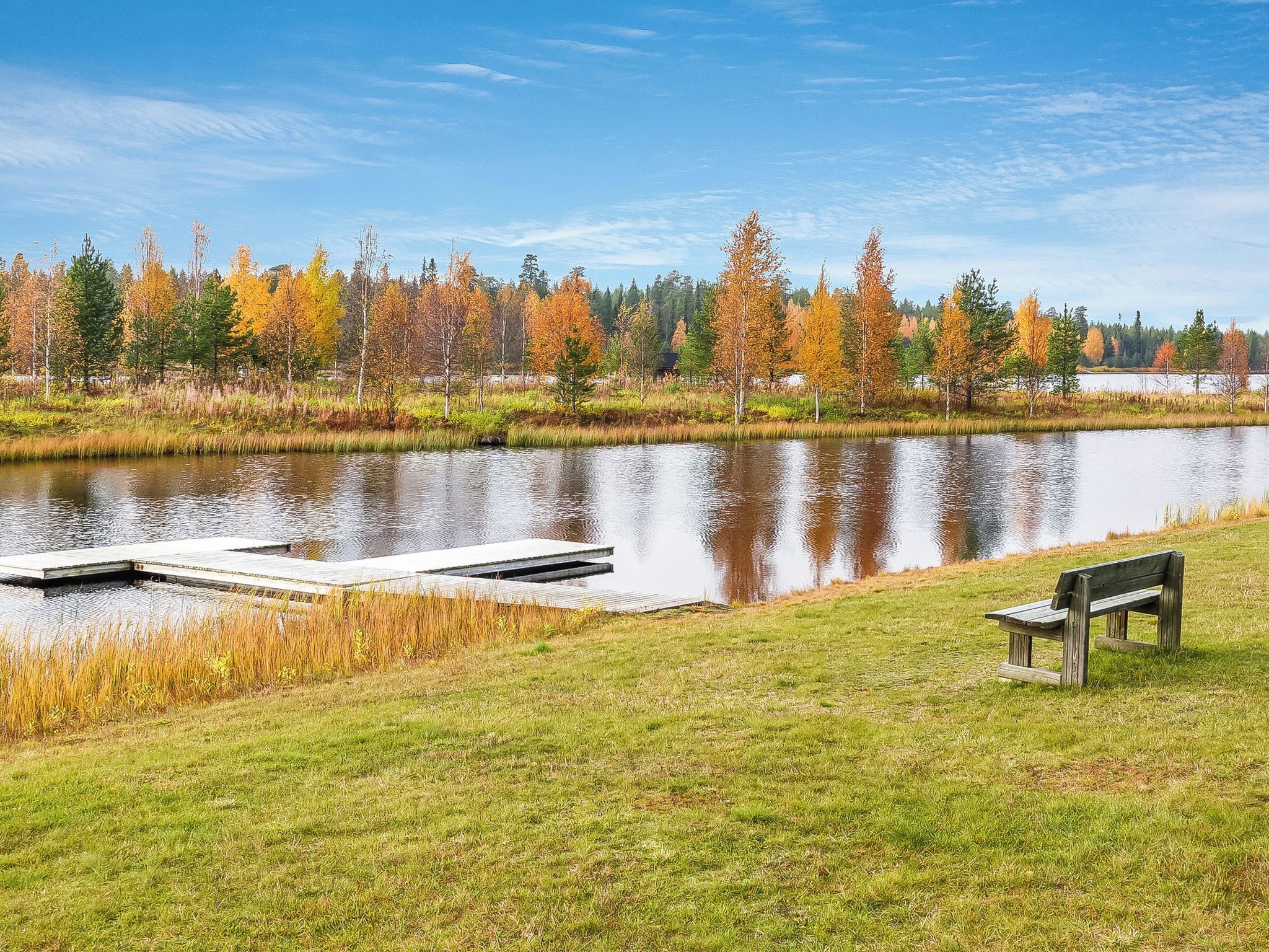 A calm lake with a dock and a bench, surrounded by trees with autumn foliage.