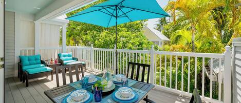 A covered outdoor deck with a table set for dining, surrounded by white railings and lush greenery.