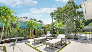 An outdoor patio with lounge chairs and a small table, surrounded by lush greenery and a fence.