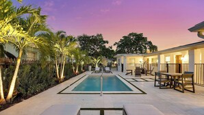 A swimming pool with lounge chairs and a covered seating area, surrounded by palm trees and a well-maintained garden.