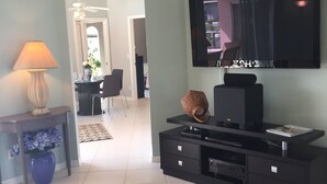 Ein Wohnzimmer mit einem Wand-Fernseher, einer schwarzen Unterhaltungseinheit mit Schubladen, einer Lampe auf einem Beistelltisch und einer blauen Vase mit Blumen.