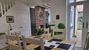 A dining area with a wooden table and white chairs, a brick wall, and a view of the garden through an open door.