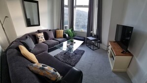 A modern living room with a dark grey sofa, a glass coffee table, a small side table, a TV stand, and a large window with curtains.