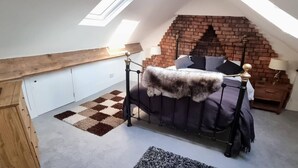 A bedroom with a sloped ceiling, a brick wall, a bed with a fur throw, a checkered rug, and a nightstand with a lamp.