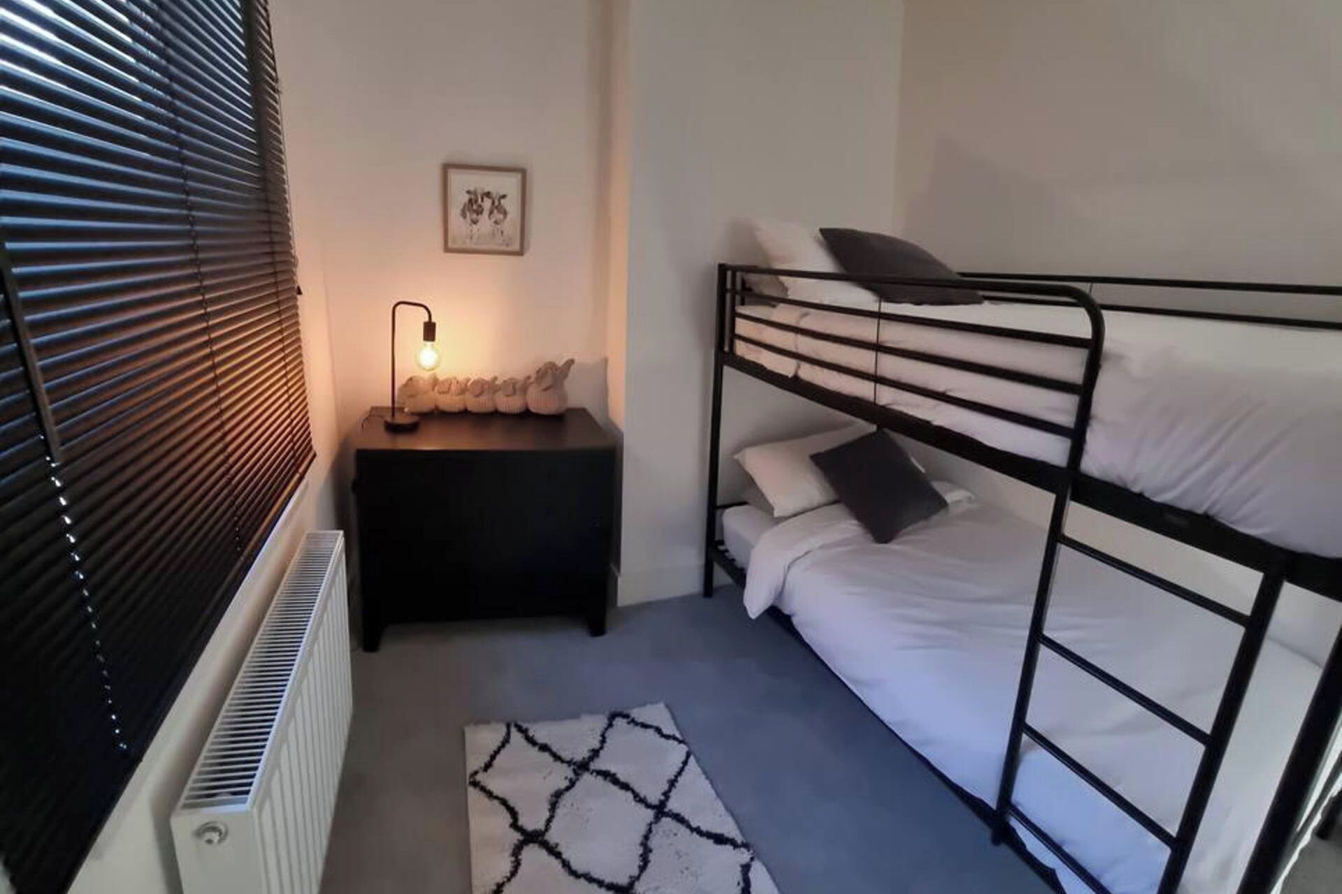 Habitació petita i senzilla amb un llit superposat, una taula de nit amb una làmpada i un radiador.