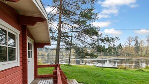 Une terrasse en bois avec une rambarde rouge mène à une maison dotée d’une porte et de fenêtres blanches. En arrière-plan, on aperçoit un lac où se reflètent les arbres, ainsi qu’un ciel dégagé.