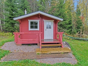 Une petite cabane rouge avec une porte et des fenêtres blanches, un porche en bois et un chemin de gravier menant à l’entrée.