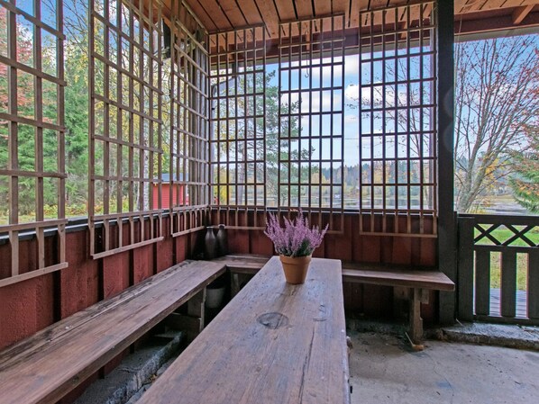 Een overdekt terras met houten banken en een tafel, met uitzicht op een weg en bomen.