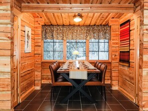 Un intérieur en bois avec une table à manger dressée pour quatre personnes, entourée de fauteuils en cuir, d’un rideau à motifs et d’une fenêtre donnant sur des arbres couverts de neige.